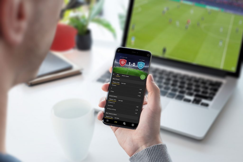 Personne tenant un smartphone avec une application de pronostics sportifs ouverte, sélectionnant un match pour parier, dans un salon confortable